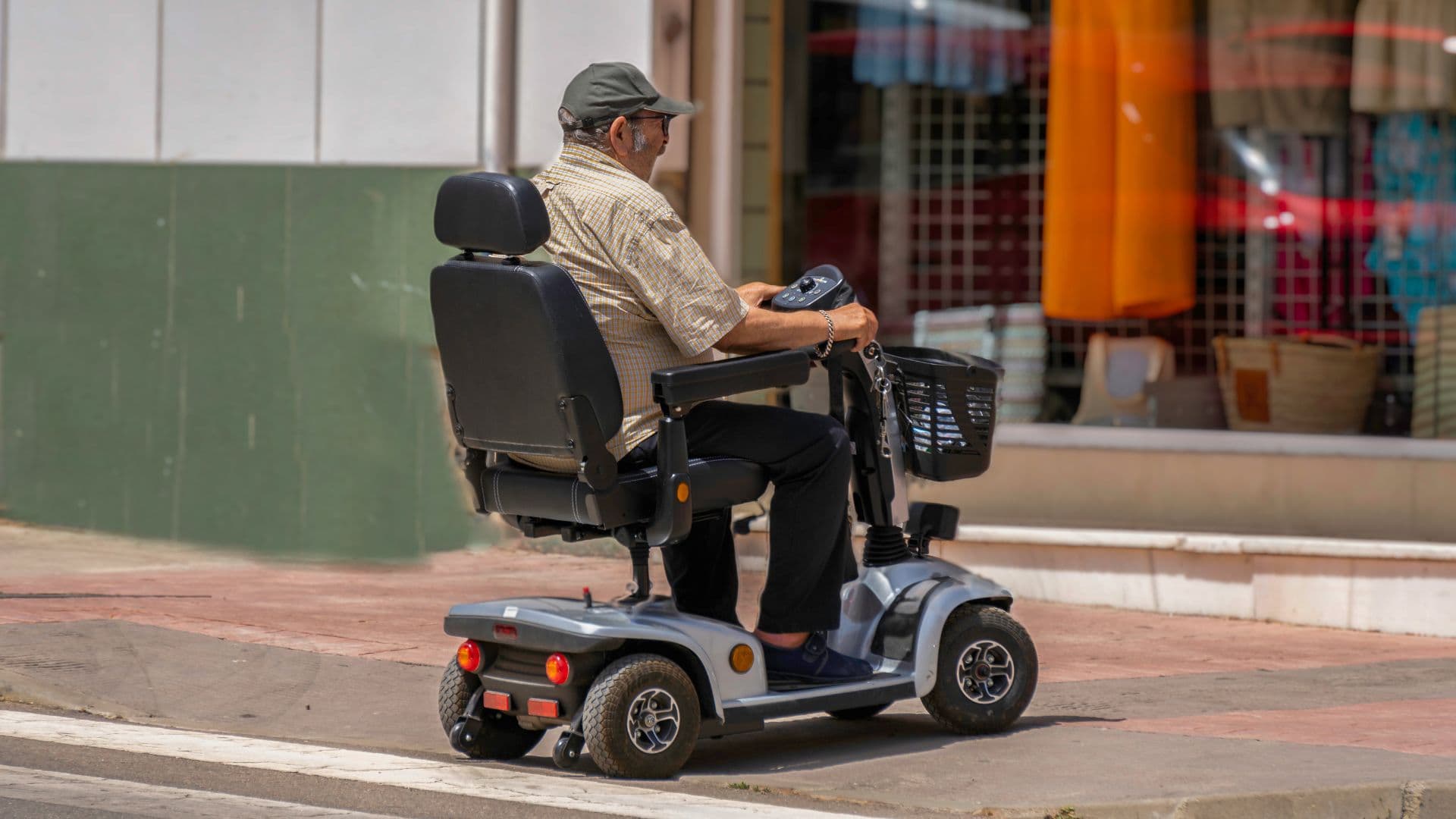 A man riding a mobility scooter along a sunlit street past a row of shopfronts