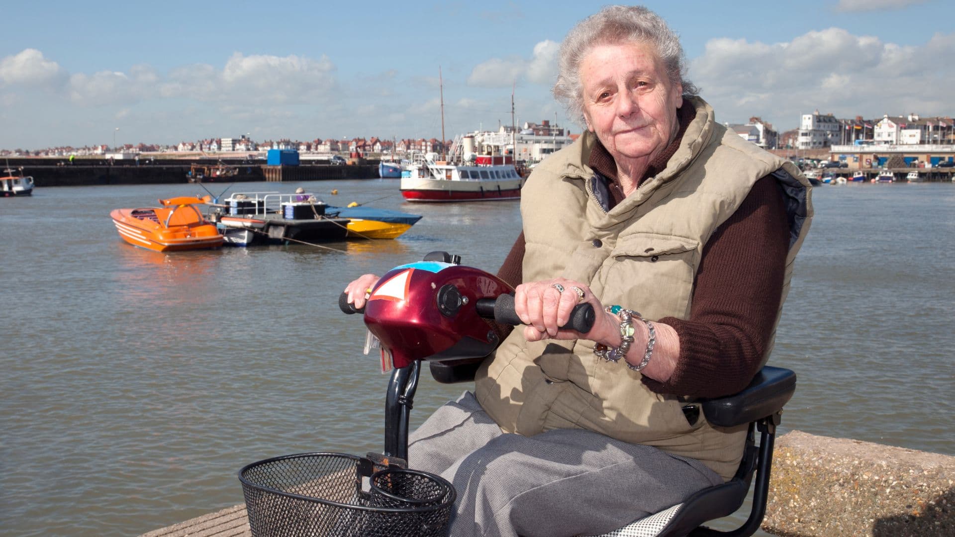 A woman on a red mobility scooter beside a harbour on a bright day